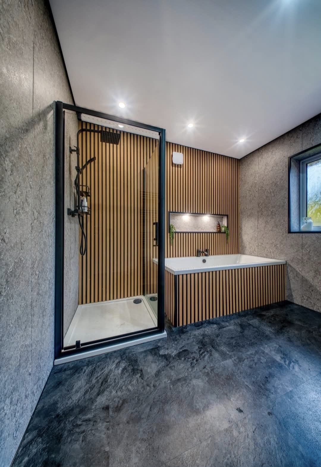 Black-framed glass shower beside an oak slat wall and corner bath