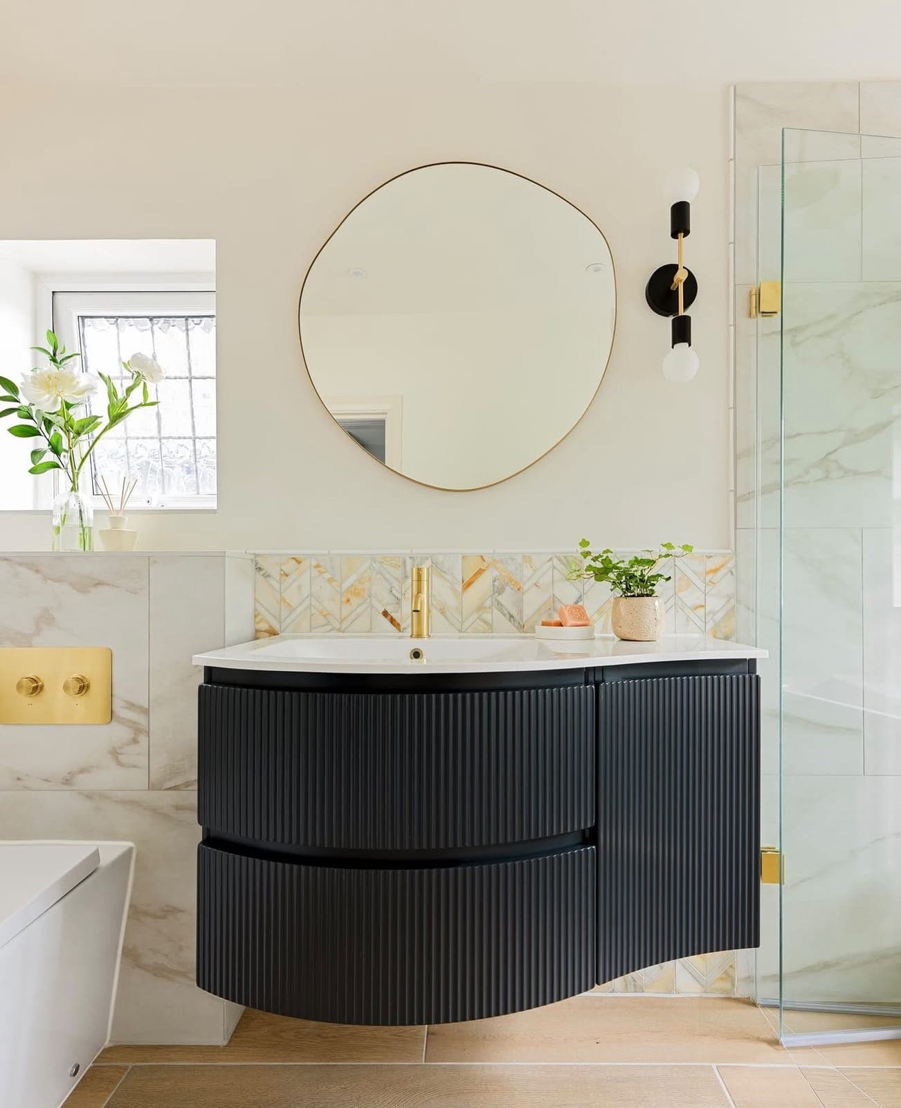 Curved black vanity with gold taps and oval mirror against marble tiles