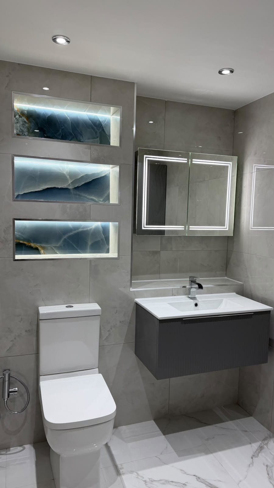 Grey tiled bathroom with dark wall-hung vanity, lit shelving niches and mirror cabinet