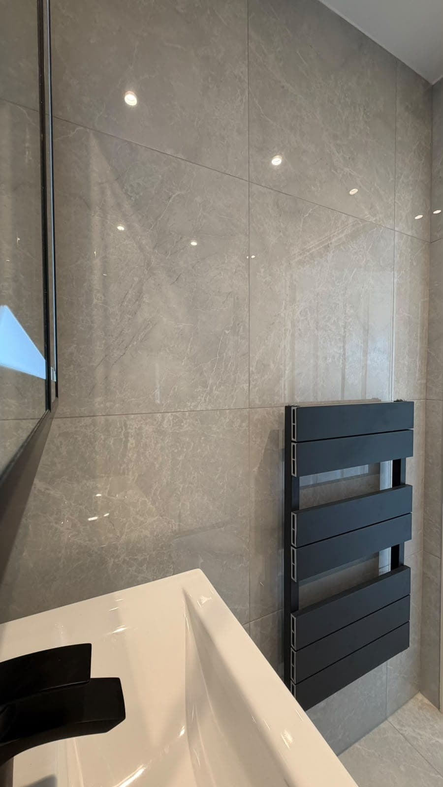 Bath and black towel radiator beside a fluted vanity in a grey-tiled bathroom
